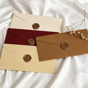 Könnte beinhalten: Vier Umschläge in Creme, Burgunderrot und Braun, versiegelt mit goldenen Wachssiegeln. Die Umschläge sind auf einer weißen Satin-Oberfläche angeordnet, mit einem Zweig weißer Blumen. Die Wachssiegel haben ein florales Design.