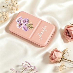 Puede incluir: Una caja metálica rectangular de color rosa claro con una ilustración impresa de flores de áster moradas en el lado izquierdo y el nombre Jennifer escrito en letra cursiva blanca en el lado derecho de la tapa.