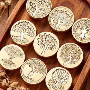 May include: A selection of gold-coloured wax seal stamps, each with a different tree design. The stamps are displayed on a wooden tray, highlighting the intricate tree illustrations. The designs feature detailed branches and foliage.