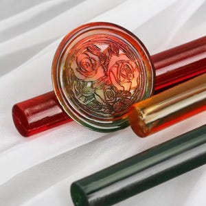 Puede incluir: Un tapón de botella decorativo de vidrio con un diseño de rosas, en tonos rojos, naranjas y verdes. Varias varillas de vidrio rojas, naranjas y verdes están en el fondo. El tapón es redondo con un patrón floral detallado.