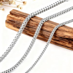 Puede incluir: Primer plano de tres collares de cadena plateados con un diseño tejido. Las cadenas se muestran sobre un trozo de madera marrón, con un fondo blanco. Los collares varían en grosor.