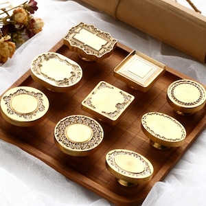 May include: A collection of ornate, gold-colored cabinet knobs in various shapes, including oval, round, and square. Each knob features intricate embossed detailing and a smooth, central surface. The knobs are displayed on a wooden tray.