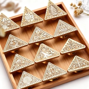 May include: A collection of twelve gold-colored, triangle-shaped cabinet knobs with various engraved designs, including leaves, crowns, and shells. The knobs are arranged in a wooden tray, showcasing their intricate details and decorative appeal.