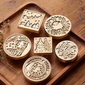 Puede incluir: Colección de seis sellos de cera de latón con varios diseños, incluyendo un paisaje, una corona y un faro. Los sellos están dispuestos sobre una bandeja de madera, con flores secas en la esquina. Los sellos tienen varias formas, incluyendo cuadrada y redonda.
