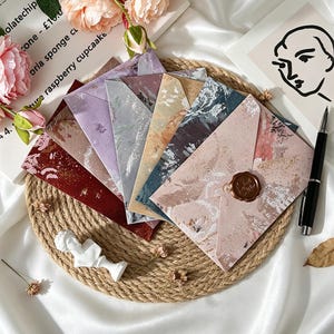 May include: A collection of overlapping envelopes in various colors including deep red, lavender, gray, tan, dark blue, and light pink, each featuring a textured, marbled pattern. One light pink envelope is sealed with a brown wax seal.