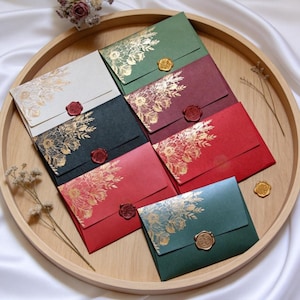 Puede incluir: Un surtido de sobres coloridos con diseños florales dorados y sellos de cera, dispuestos en una bandeja de madera. Los sobres son en tonos de rojo, verde y crema, con detalles dorados. Un elegante juego de papelería.