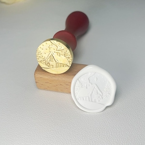 Könnte beinhalten: Ein Messing-Wachssiegelstempel mit Holzgriff und Winterhütten-Design. Ein weißes Wachssiegel mit dem gleichen Hüttendesign befindet sich neben dem Stempel. Der Stempel befindet sich auf einem kleinen Holzblock.