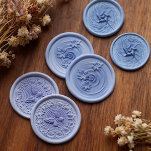 Puede incluir: Una colección de sellos de cera azul claro con varios diseños en relieve. Algunos sellos presentan patrones florales, mientras que otros muestran una mariposa o un diseño de molinillo. Los sellos están dispuestos sobre una superficie de madera, con flores secas en el fondo.