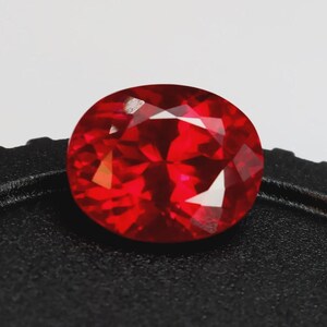 Puede incluir: Una gema roja brillante, facetada y de forma ovalada. La gema tiene múltiples superficies reflectantes, creando una exhibición brillante de luz. La piedra se encuentra sobre un fondo oscuro y texturizado, resaltando su color vibrante y claridad. Esta gema podría usarse en joyería.