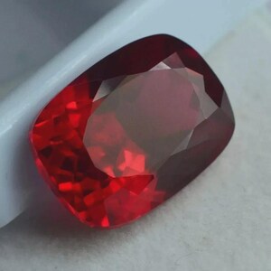 May include: A faceted, cushion-cut gemstone in a deep red colour. The gem is set against a white background, highlighting its brilliance. The cut of the stone allows light to reflect, creating a sparkling effect.