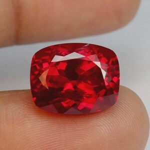 May include: A faceted, cushion-cut, deep red gemstone. The gem is held between fingers, showcasing its brilliant, reflective surfaces. The stone's color ranges from a vibrant, bright red to a darker, more intense hue, with sharp, defined edges.