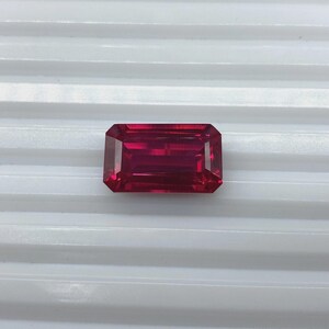 Può includere: Una gemma rettangolare e sfaccettata di colore rosso intenso. La gemma ha un taglio pulito e simmetrico, con una serie di sfaccettature parallele che creano un brillante gioco di luce. La gemma è posta su uno sfondo bianco.
