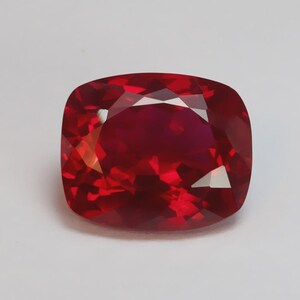 Peut inclure: Un rubis taillé en coussin, d'un rouge profond et intense. La surface à facettes de la pierre précieuse reflète la lumière, créant un effet scintillant. La forme est un carré arrondi, avec une finition polie. Le rubis est posé sur un fond blanc.