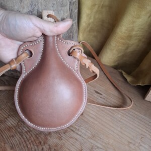 May include: A round, tan leather canteen with a smooth, molded front panel and visible white stitching along the edges. The canteen features a wooden stopper at the top and includes a long, thin leather strap attached to two side loops.