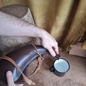May include: A dark brown leather canteen with a curved shape and light tan leather straps is pouring clear liquid into a small black metal mug with a white interior. The canteen features visible stitching along its edges and a narrow spout.
