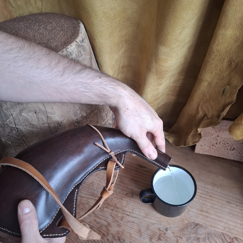 May include: A dark brown leather canteen with a curved shape and light tan leather straps is pouring clear liquid into a small black metal mug with a white interior. The canteen features visible stitching along its edges and a narrow spout.