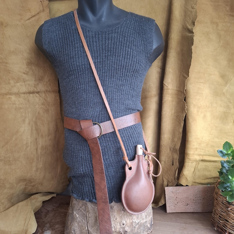 May include: A dark gray sleeveless ribbed top worn with a wide brown leather belt featuring a metal ring buckle. A brown leather canteen with a light-colored wooden stopper is suspended from a long, thin leather strap worn across the torso.