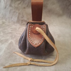 Può includere: Un piccolo sacchetto di pelle marrone con chiusura a coulisse. La borsa ha un lembo decorativo in pelle con cuciture bianche e un emblema circolare. Un cinturino in pelle è attaccato in alto per appenderlo.