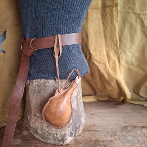 Puede incluir: Un cinturón de cuero marrón con una hebilla de anillo metálico y una pequeña petaca de cuero en forma de pera sujeta por un cordón de cuero fino. El cinturón rodea una prenda acanalada gris oscuro y la petaca cuelga sobre una base de madera.