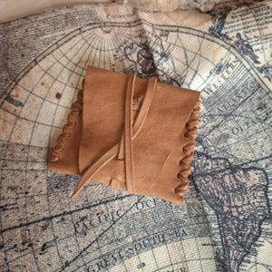 May include: A small, square, tan leather pouch featuring visible whip-stitched edges along the sides. The pouch is secured with a thin leather cord that wraps around the centre, holding the folded flap in place. The surface of the leather is smooth.