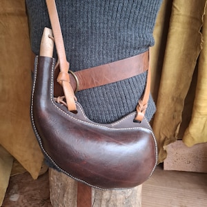 May include: A dark brown leather canteen with a light-colored wooden stopper, featuring white stitching along the edges. The canteen is suspended from a tan leather strap that is attached to a wider brown leather belt worn around the waist.