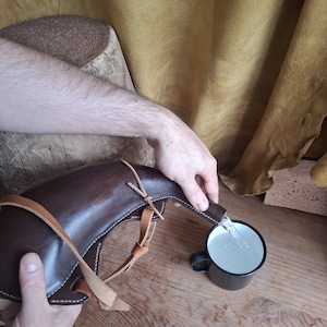 May include: A dark brown leather canteen with light tan leather straps and stitching, pouring clear liquid into a small black metal mug. The canteen has a curved, horn-like shape and is held by two hands, showcasing the functional design.