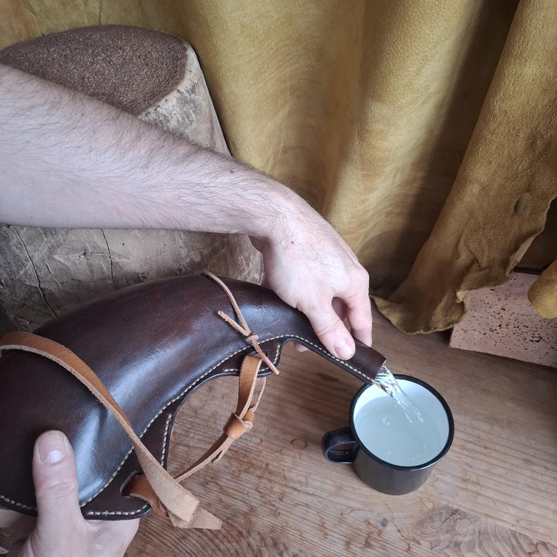 May include: A dark brown leather canteen with light tan leather straps and stitching, pouring clear liquid into a small black metal mug. The canteen has a curved, horn-like shape and is held by two hands, showcasing the functional design.