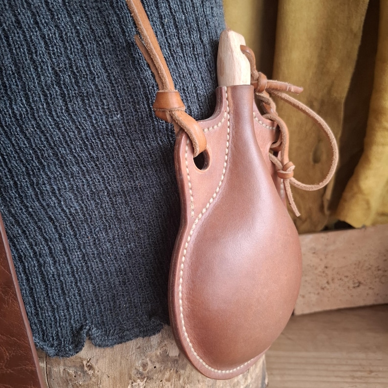 May include: A brown leather canteen with a rounded body and a light-colored wooden stopper. The canteen features visible white stitching along the edges and is attached to a matching leather strap for carrying, showcasing a smooth, firm texture.