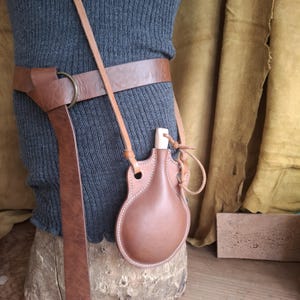 May include: A brown leather canteen with a light-colored wooden stopper, suspended by a thin leather strap from a wide brown leather belt featuring a brass ring buckle. The canteen has a rounded, flask-like shape with visible stitching along the edges.