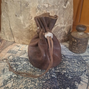 May include: A brown leather pouch with a drawstring closure. The pouch is gathered at the top and secured with a light-colored, decorative clasp. The pouch is approximately 4 inches tall.
