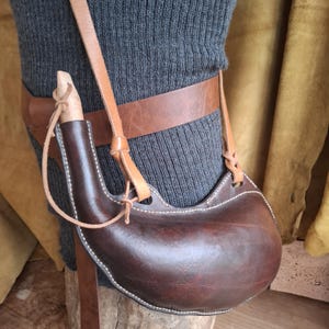 May include: A dark brown leather canteen with a curved shape and white stitching, featuring a light-colored wooden stopper and a tan leather shoulder strap. The canteen is attached to a wide, smooth brown leather belt worn around the waist.
