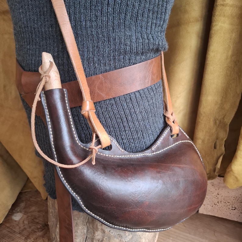 May include: A dark brown leather canteen with a curved shape and white stitching, featuring a light-colored wooden stopper and a tan leather shoulder strap. The canteen is attached to a wide, smooth brown leather belt worn around the waist.