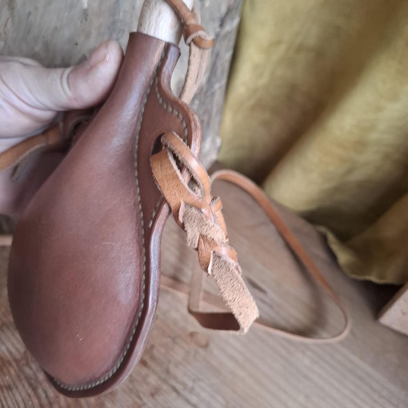 May include: A brown leather canteen with a rounded body and a wooden stopper. The canteen features stitched edges and a braided leather strap attached to the side, designed for carrying. The leather has a smooth, matte finish with visible seams.