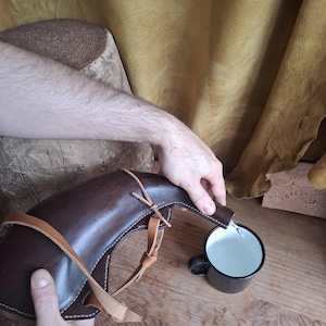May include: A dark brown leather wineskin with a curved spout and light tan leather straps pouring clear liquid into a black enamel mug. The wineskin features visible stitching along the edges and a smooth, polished surface texture.