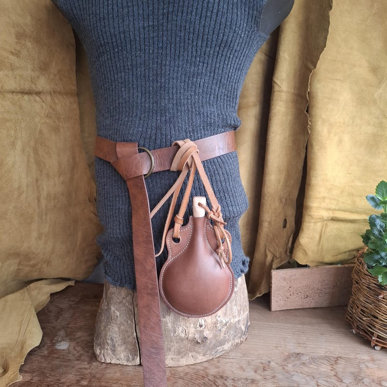 May include: A brown leather belt with a metal ring buckle and a matching brown leather canteen flask attached by thin leather cords. The canteen features a light-colored wooden stopper and is suspended from the belt, which is worn over a gray ribbed top.