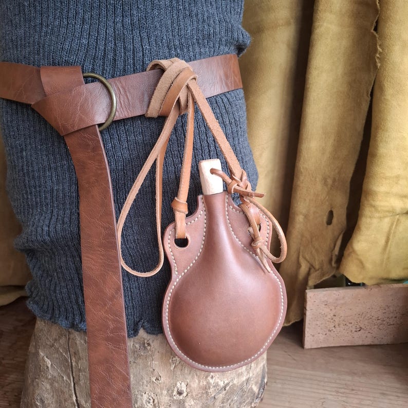 May include: A brown leather canteen with a light-colored wooden stopper, suspended by thin leather cords from a matching brown leather belt featuring a circular metal ring. The canteen has a rounded, flask-like shape with visible stitching.