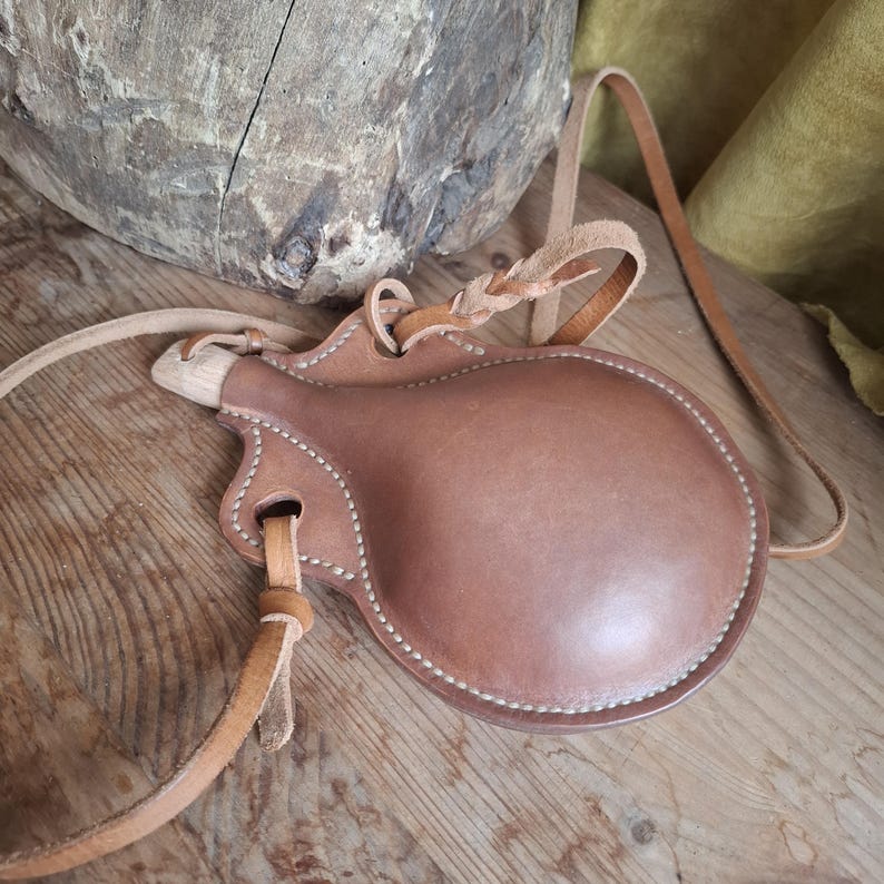 May include: A round, brown leather canteen featuring visible light-colored stitching along the edges. The canteen includes a wooden stopper at the top and a long, thin leather strap attached through braided leather loops for carrying purposes.