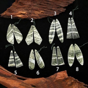 May include: Eight pairs of green and white patterned stone pendants labelled one through eight. Each pair features unique, elongated shapes with natural mineral banding and speckled textures, suspended by thin dark green threads against a black background.
