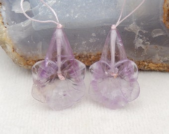 May include: Two carved amethyst flower pendants with light pink string loops. The pendants are a light purple color with translucent petals and a detailed floral design. The background is a light blue and white stone.