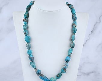 Puede incluir: Un collar de piedras turquesas expuesto sobre un maniquí blanco. El collar presenta piedras de turquesa pulidas de forma irregular, en tonos azules y marrones. Las piedras están ensartadas para formar un collar.