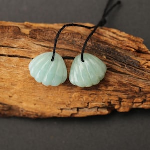May include: Two light blue, seashell-shaped pendants with black cords. The pendants have a ribbed texture and are displayed on a piece of weathered wood. The background is a dark gray.