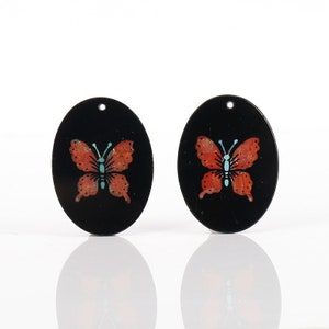 May include: Two black oval-shaped pendants with a red butterfly design. The butterflies have blue accents on the wings.
