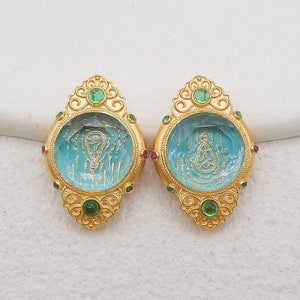May include: A pair of ornate gold earrings. Each earring features a light blue, oval-shaped center with a gold design. The earrings are adorned with green and red gemstones. The gold setting has a decorative, filigree design.