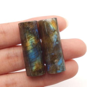 Puede incluir: Dos piedras preciosas de labradorita de forma cilíndrica con una superficie pulida que muestra destellos iridiscentes azules y dorados sobre una base marrón oscura. Estas piedras presentan inclusiones minerales naturales y un acabado liso.