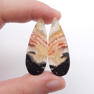 Puede incluir: Dos colgantes de piedras preciosas en forma de lágrima. Las piedras presentan una mezcla de colores, incluyendo negro, crema, naranja y rojo, con un patrón veteado. Cada colgante tiene un pequeño agujero en la parte superior para ensartar.