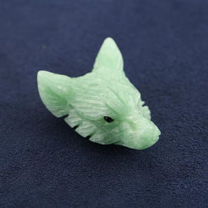 Könnte beinhalten: Eine hellgrüne, geschnitzte Steinstatuette in Form eines Fuchskopfes. Die Skulptur zeigt detaillierte Fellstrukturen, spitze Ohren und eine kleine, dunkle, kreisförmige Einlage für das Auge, die die Gesichtszüge des Tieres glatt und poliert darstellt.