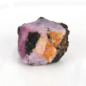 May include: A rough, pink and orange gemstone with black inclusions. The gemstone is a raw, unpolished crystal.