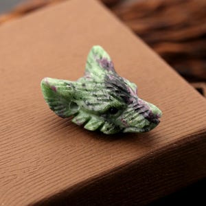 May include: A carved stone wolf head bead featuring a mottled green and pink color palette with dark black inclusions. The detailed carving shows pointed ears, a snout, and a small circular hole on the side for threading onto a cord or wire.