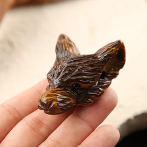 May include: A carved tiger's eye gemstone shaped into the head of a fox or wolf. The stone features a polished, chatoyant surface with deep golden-brown and dark brown bands, showing detailed fur textures and dark, smooth, rounded eyes.