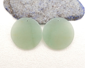 Può includere: Due pendenti rotondi in pietra preziosa verde pallido, con piccoli fori per l'infilatura. I pendenti sono lisci e traslucidi, con una leggera variazione di colore. Sono esposti su una superficie bianca con una pietra strutturata grigia e blu sullo sfondo.